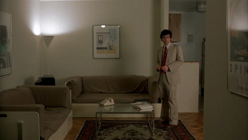 After Hours (1985) movie still: fullbody — A spare, furnished apartment room features two couches, a coffee table, and beige walls. A man in a tan…