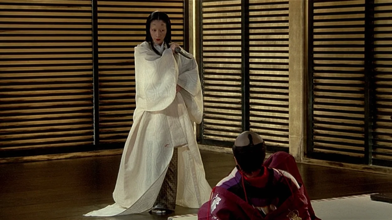 Ran (1985) movie still: fullbody — A room features dark wood flooring and window panels with horizontal wooden slats. A woman in a white robe stands…