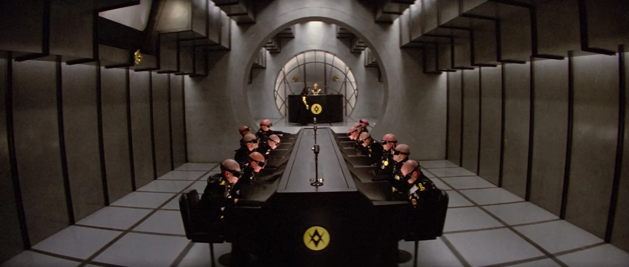 Flash Gordon (1980) movie still: wide — A sterile, gray, brutalist meeting room features a large circular archway and grid flooring. Twelve…