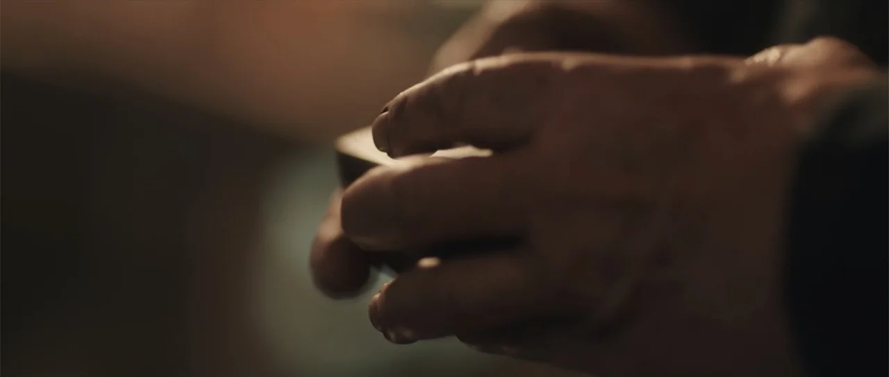 Night of the Missing (2023) movie still: closeup — A close view of adult hands holding a small dark object in a dimly lit space. The fingers are…
