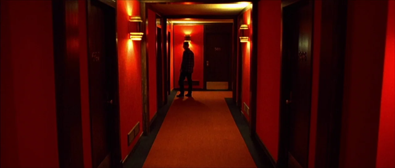 Fear X (2003) movie still: wide — A long, symmetrical hallway features intense red-painted walls and a matching red runner rug. A man stands in the…