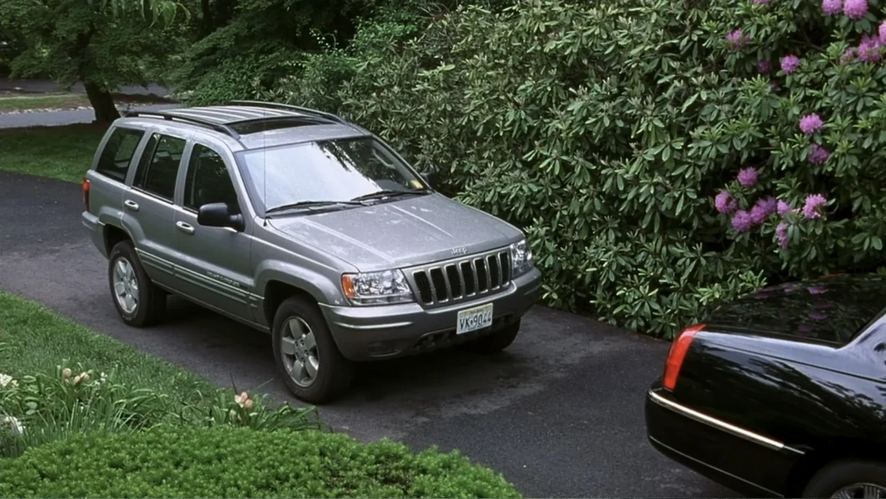 Analyze That (2002) movie still: wide — A silver SUV and a black sedan are parked in a driveway. The driveway is bordered by dense green bushes…