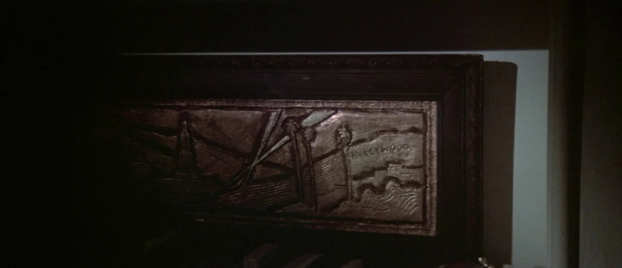 The Long Goodbye (1973) movie still: closeup — A relief-carved bronze plaque hangs on a plain wall in a dimly lit space. The artwork features an…