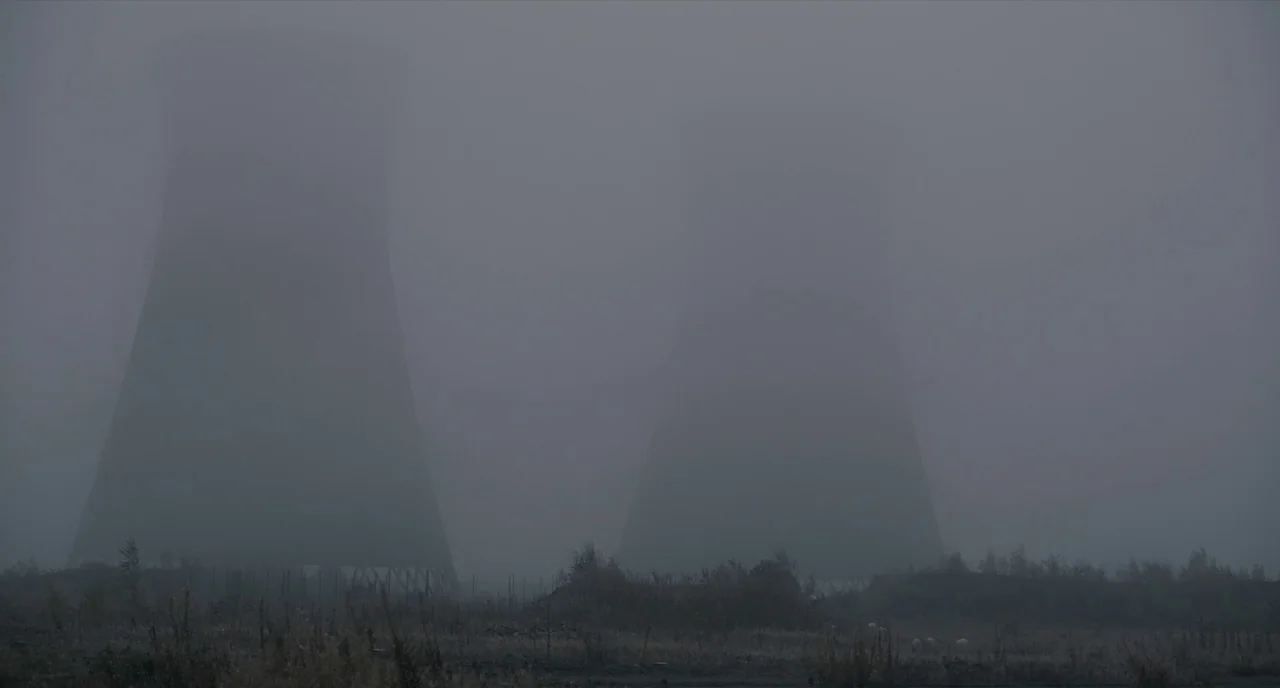 The Selfish Giant (2013) movie still: establishing — Two massive industrial cooling towers rise above a dry, grassy field in thick fog. The…