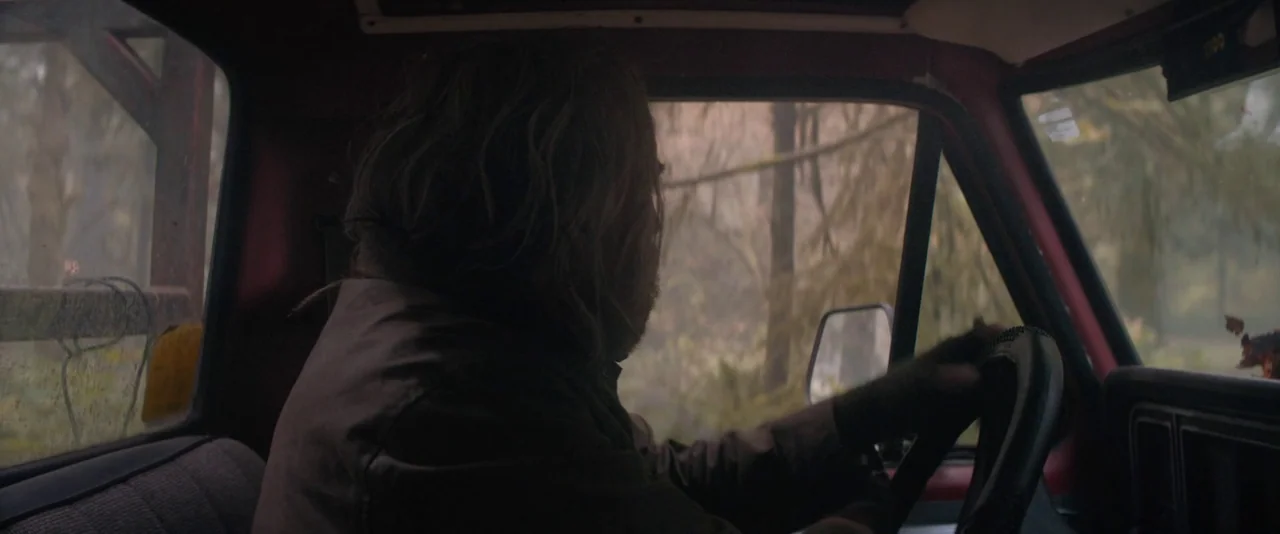 Pig (2021) movie still: medium — The interior of an older truck with a red painted frame and dark fabric seating. A man with long brown hair drives…