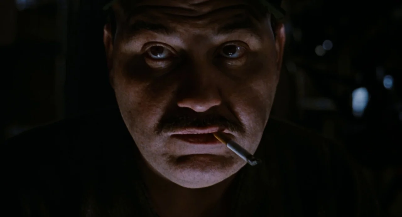 The Crow (1994) movie still: closeup — A dimly lit room with deep shadows obscuring the background. A man with a mustache looks forward while…