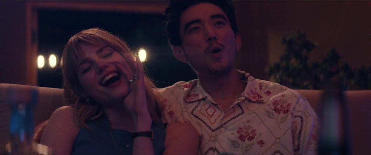 The Greatest Hits (2024) movie still: medium — A dimly lit room at night provides a muted backdrop for two people sitting together. The woman laughs…