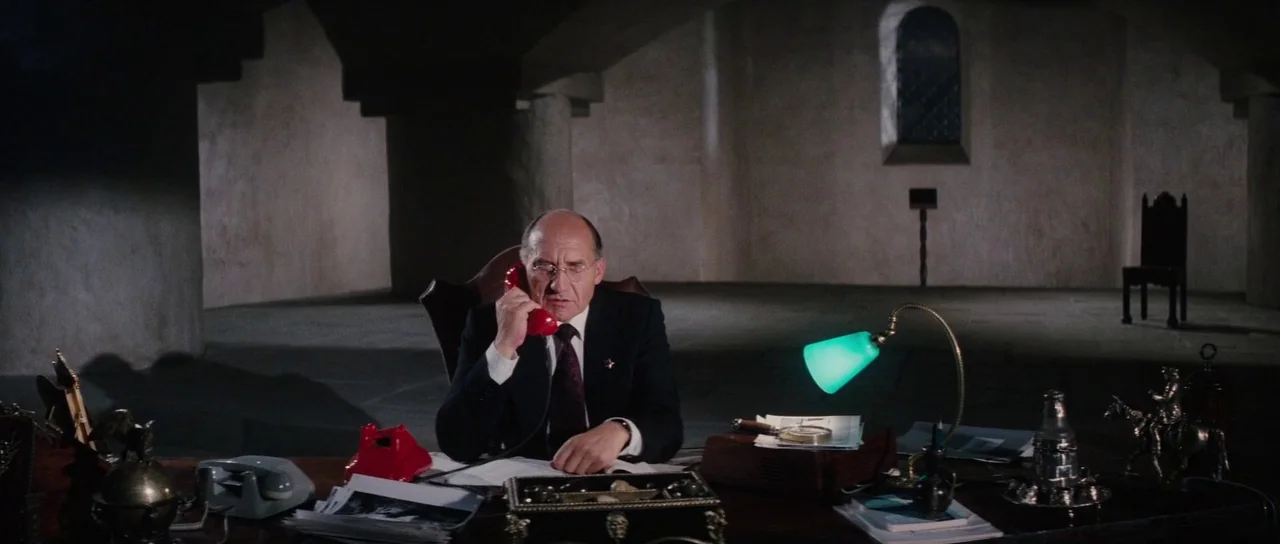 The Spy Who Loved Me (1977) movie still: medium — A dark, sparsely furnished room with arched stone walls and a single stone window. A man sits at a…