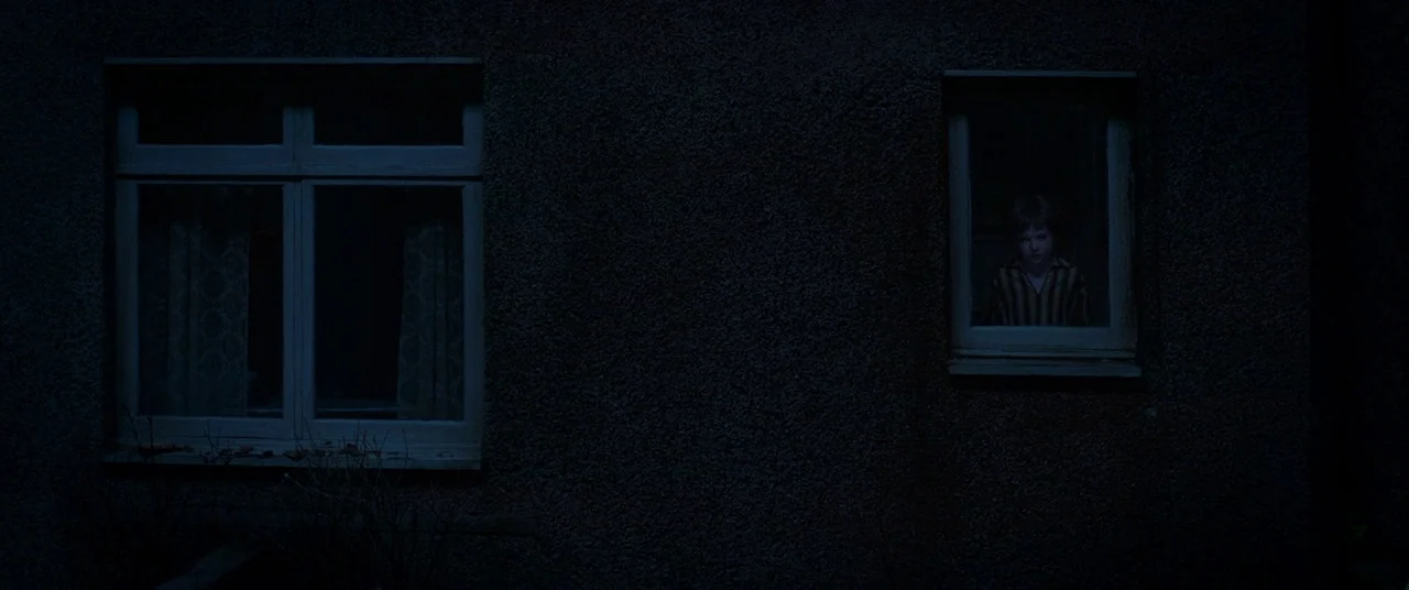 The Conjuring 2 (2016) movie still: wide — The exterior of a house at night features dark, textured stone walls and two windows. A small boy stands…