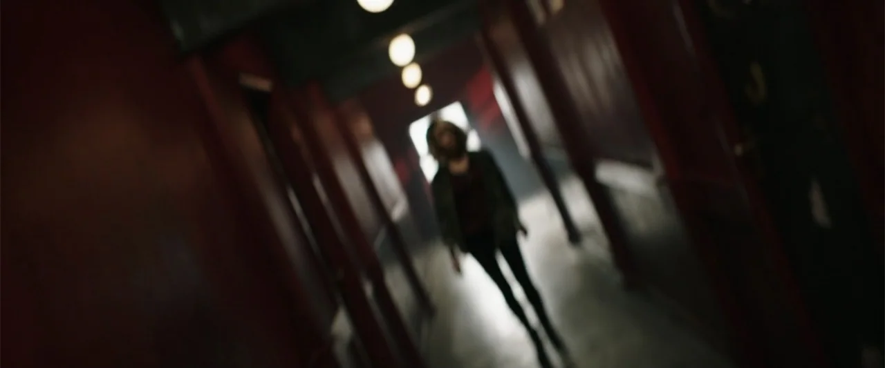 Jolt (2021) movie still: medium — A dark, narrow corridor with red-painted walls and a series of hanging overhead lights. A person walks toward a…