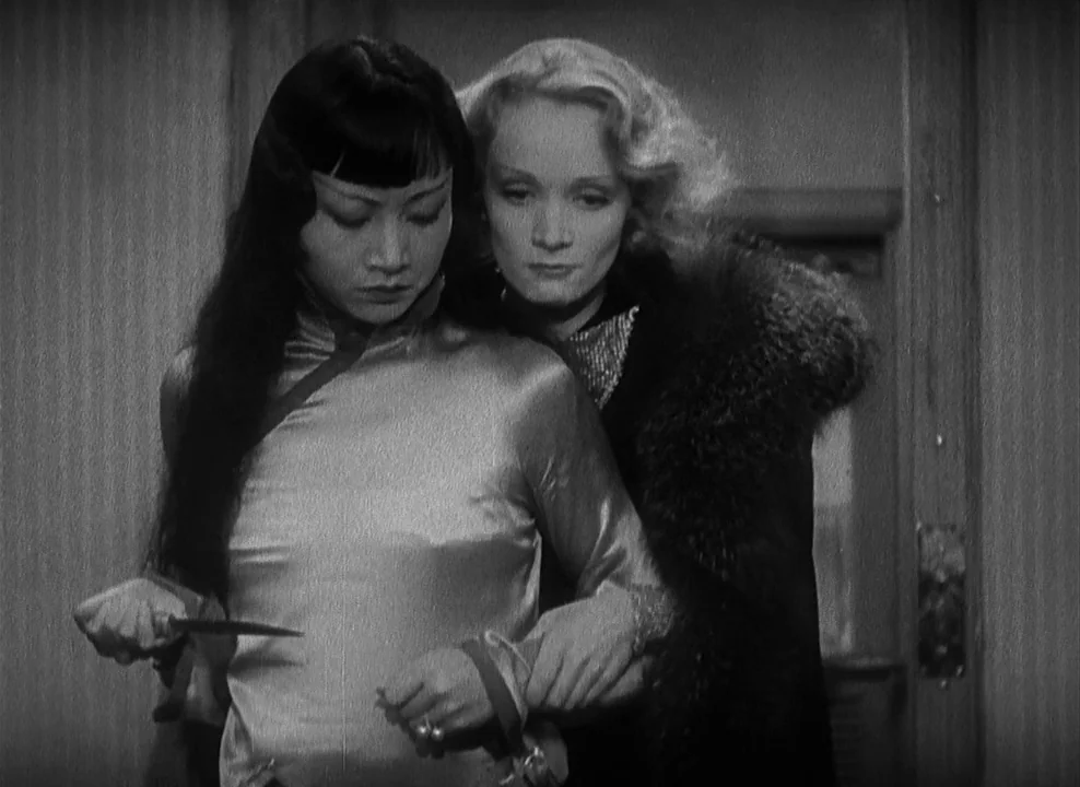Shanghai Express (1932) movie still: medium — Two women stand close together inside a room with vertical paneled wooden walls. One woman holds a…