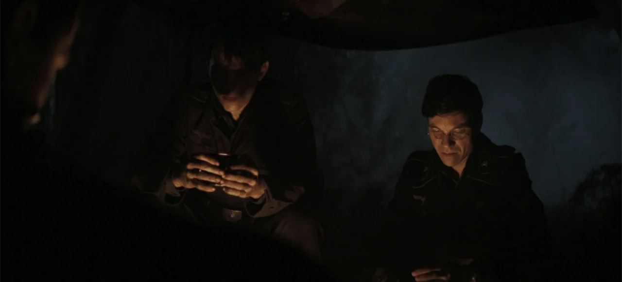 The Tank (2025) movie still: medium — Two men in dark military uniforms sit together near a fire at night. They focus intently on objects held in…