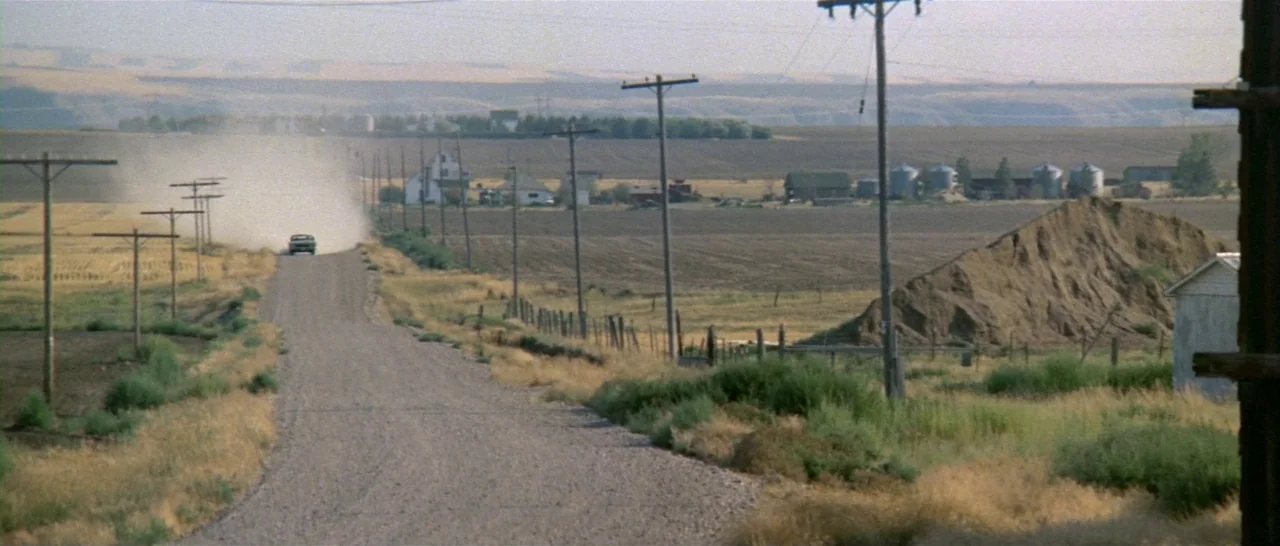 Thunderbolt & Lightfoot (1974) movie still: establishing — A rural landscape features a long gravel road extending through arid farmland fields. A…