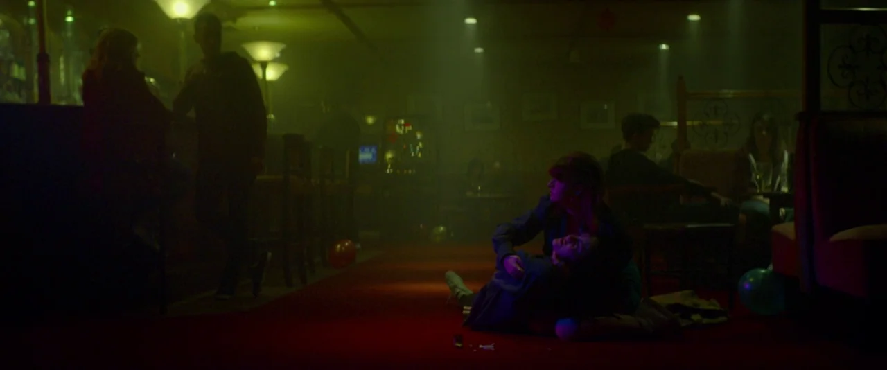Kissing Candice (2017) movie still: wide — A dimly lit, green-tinted pub interior with red carpet and blurred background patrons. A woman kneels on…