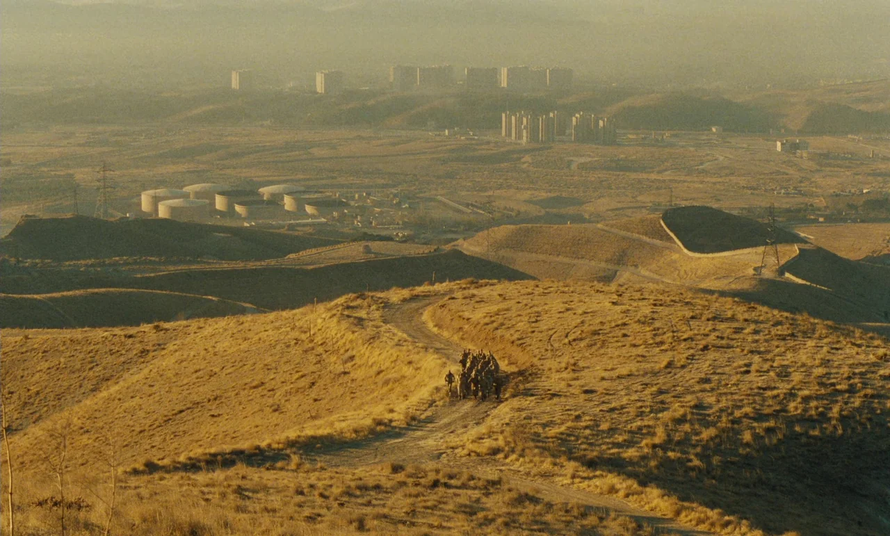 Taste of Cherry (1997) movie still: establishing — A vast, arid hilly landscape features a dirt path leading toward a distant city. A small group of…