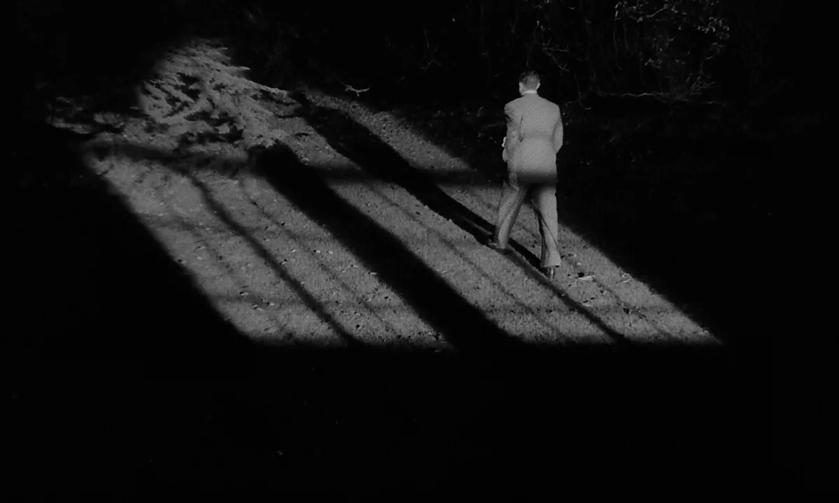 Night Of The Demon (1957) movie still: wide — A dark, unpaved path at night illuminated by a harsh, single directional light source. A man in a suit…