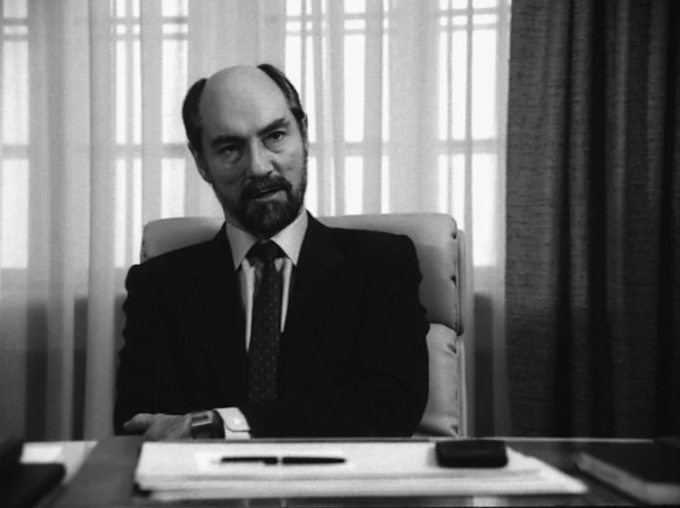 From the Life of the Marionettes (1980) movie still: medium — An office features a desk with papers and curtains covering a window background. A man…