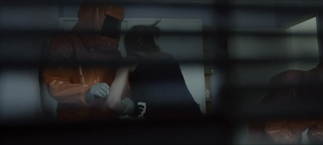 Containment (2015) movie still: medium — An interior space viewed through horizontal blinds, showing a person in an orange hazmat suit. The hazmat…