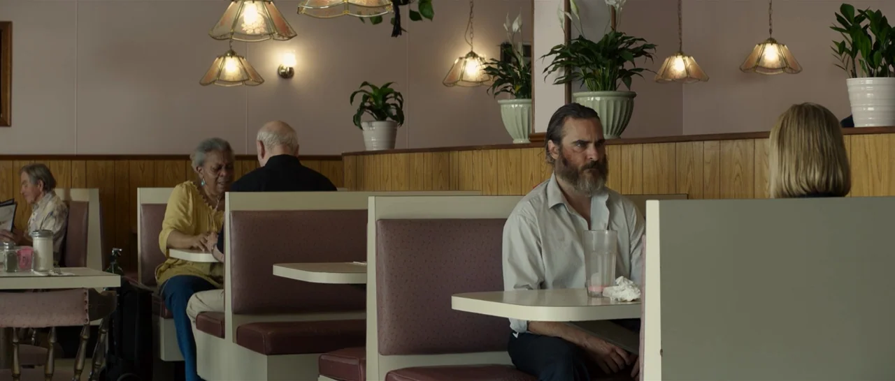 You Were Never Really Here (2017) movie still: medium — A diner booth with wood-paneled walls and potted plants on a high shelf. A man sits at a…