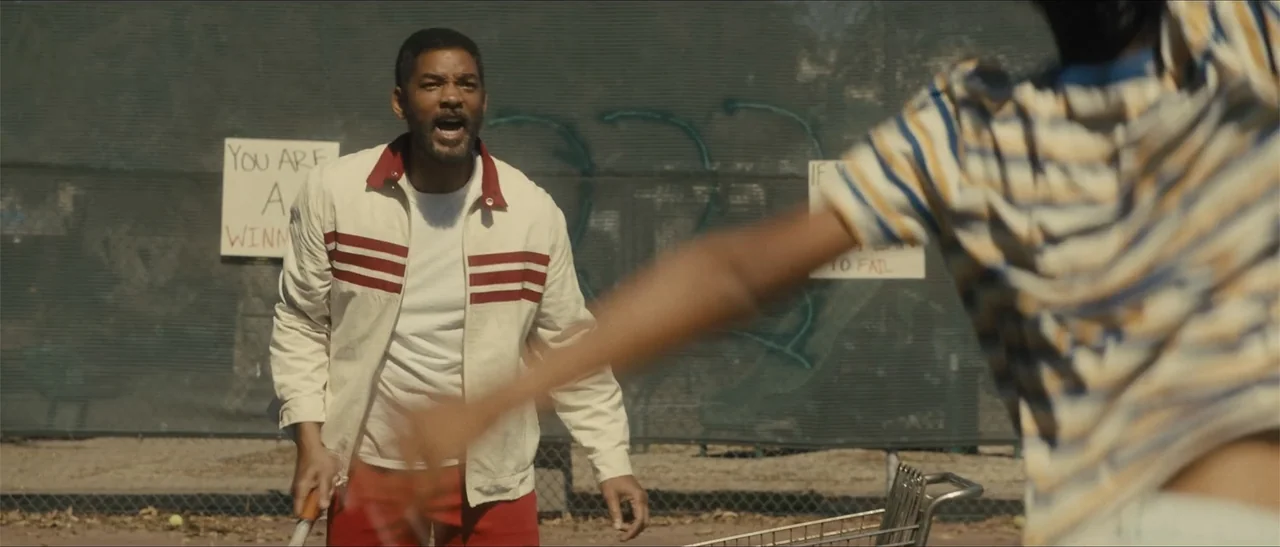 King Richard (2021) movie still: medium — An outdoor tennis court features a chain link fence with taped paper signs visible. A middle-aged man…