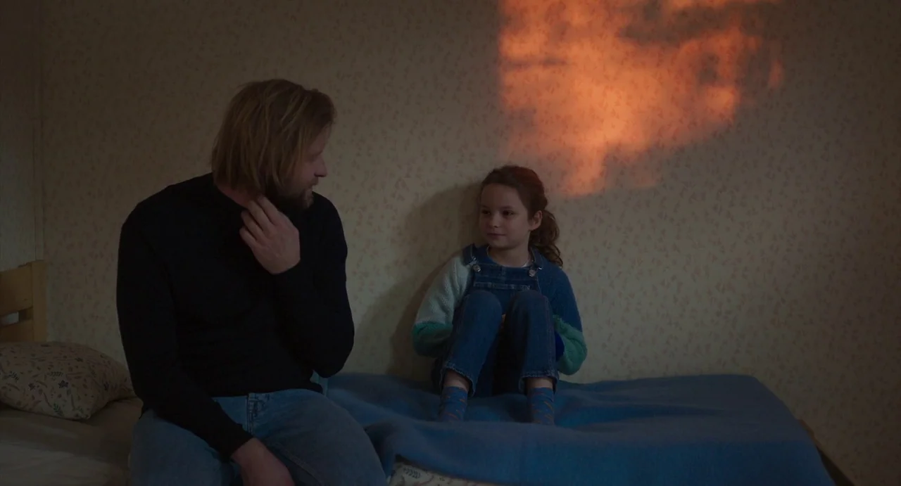 Petite Maman (2021) movie still: medium — A small room features patterned wallpaper and a blue blanket on a bed. A man and a young girl sit facing…