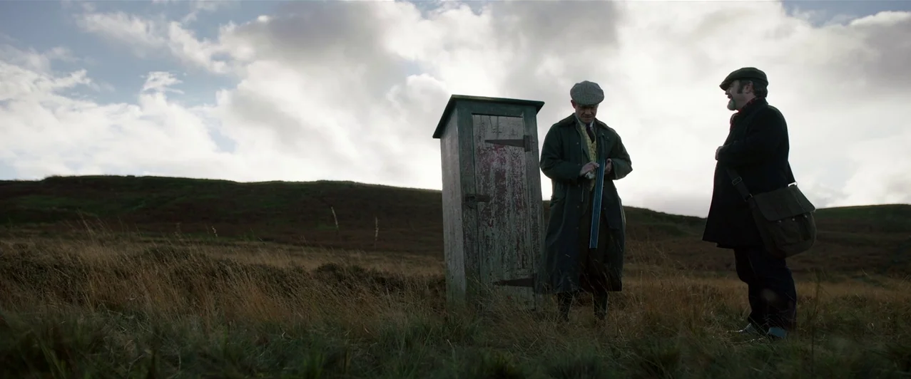 Ghost Stories (2017) movie still: wide — An open expanse of brown grassland sits under a cloudy, bright sky. Two men stand beside a weathered,…