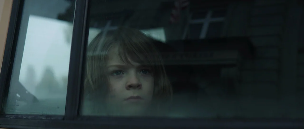 Pete’s Dragon (2016) movie still: closeup — A child peers through the window of a car in a muted, gray environment. The child looks outward with a…