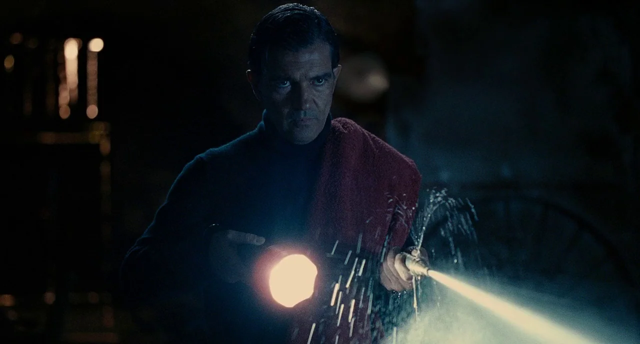 The Skin I Live In (2011) movie still: medium — A dark, dimly lit indoor space with indistinct shadows in the background. A man holds a flashlight…