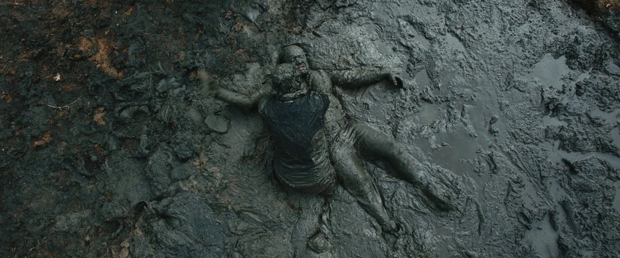 Vincent Must Die (2023) movie still: wide — A vast expanse of thick, wet, churned mud covers the entire landscape frame. Two individuals struggle…