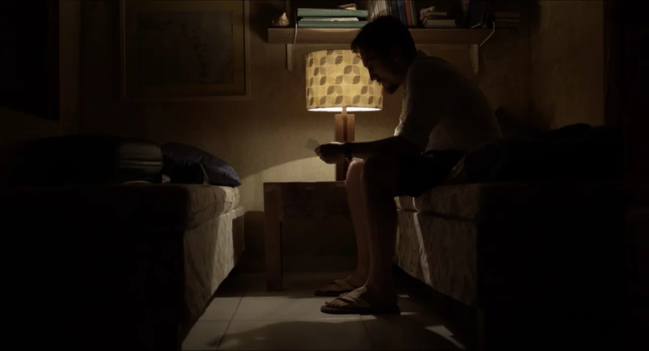 Suspension Points (2022) movie still: medium — A dark hotel room at night is illuminated by a single warm lamp. A man sits on the edge of the bed…