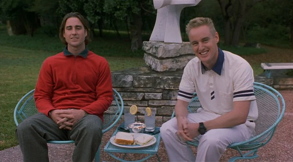 Bottle Rocket (1996) movie still: medium — Two men sit outside on a paved area near a stone sculpture. A small table between them holds glasses of…