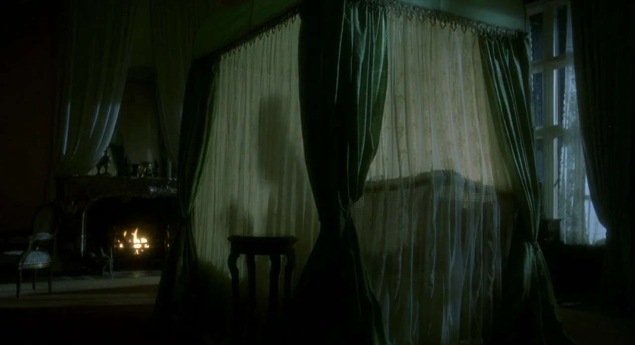 Quills (2000) movie still: wide — A dimly lit room features a draped canopy bed in the center frame. A glowing fireplace sits to the left and a…