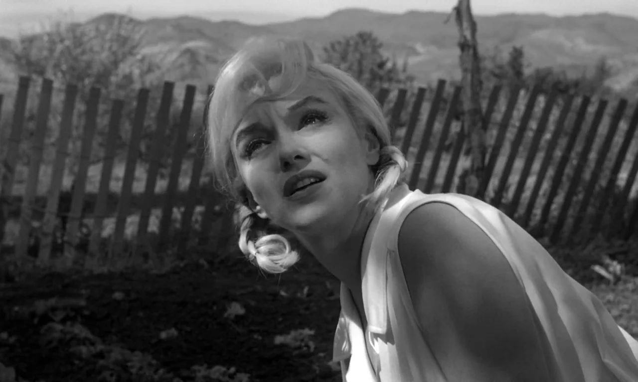 The Misfits (1961) movie still: medium — A rural landscape features rolling hills and a rustic wooden fence in the distance. A woman with blonde…