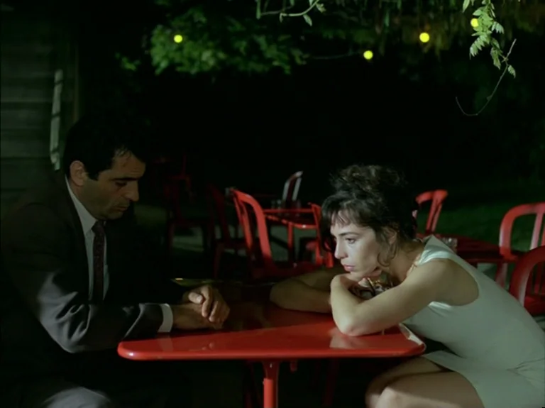 Helas Pour Moi (1993) movie still: medium — An outdoor night setting featuring a red table and several matching red chairs. A man in a suit and a…