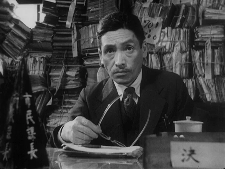 Ikiru (1952) movie still: medium — A cluttered office filled with towering stacks of paperwork and filing boxes. An official sits behind a desk…