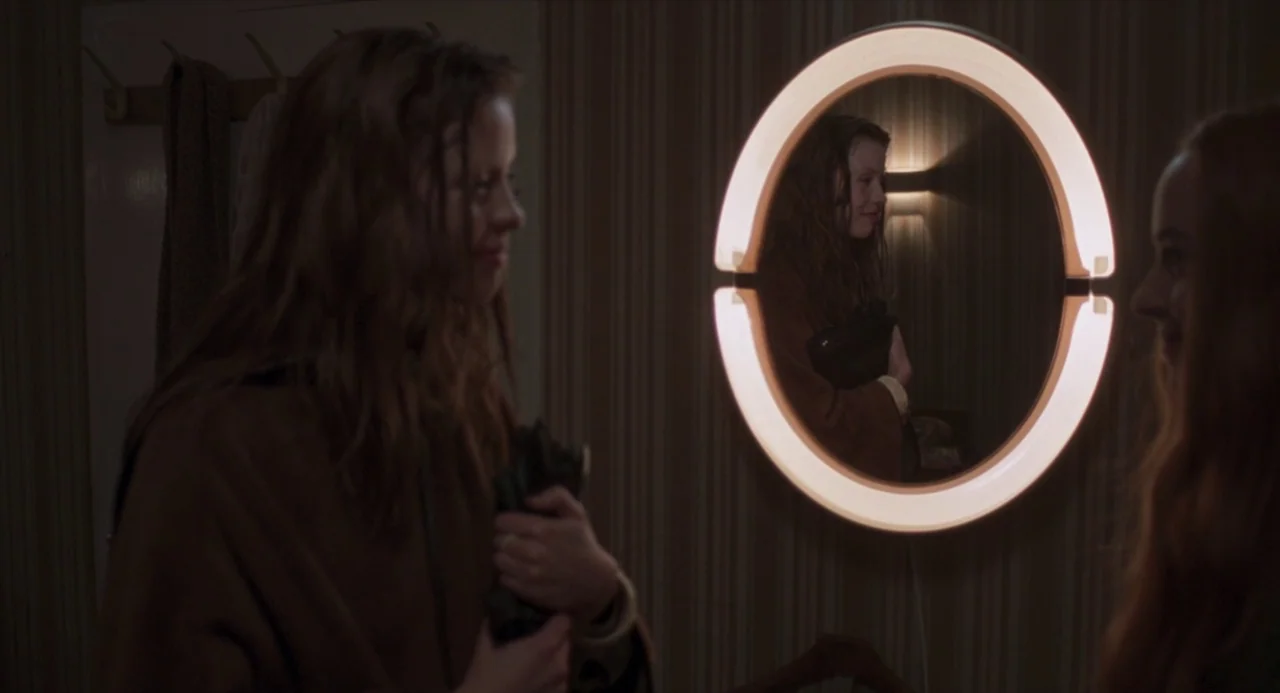 Suspiria (2018) movie still: medium — A woman stands in a dimly lit hallway with vertical-striped wallpaper. She looks into an illuminated oval…