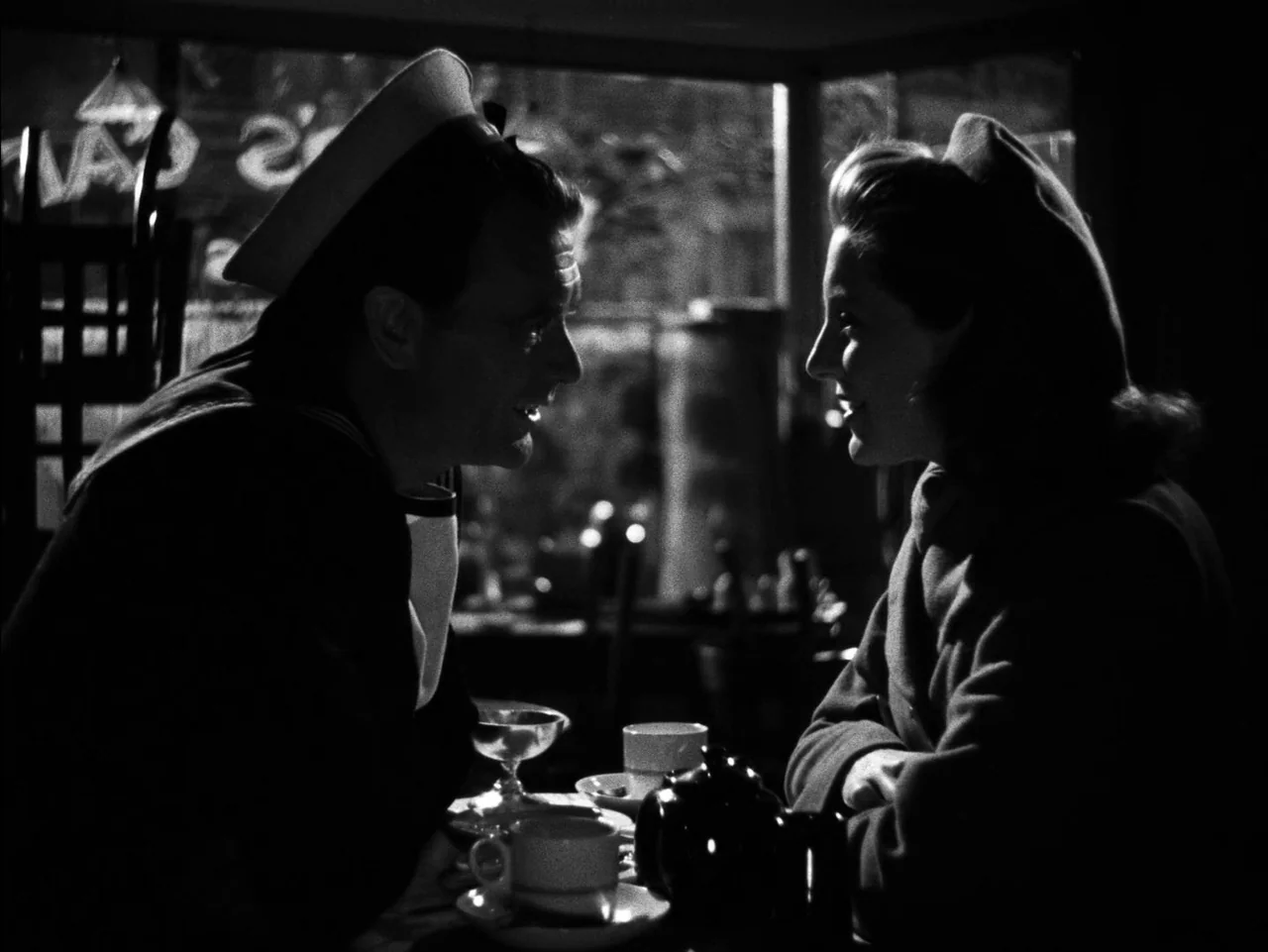 In Which We Serve (1942) movie still: medium — A dark, shadowed diner booth with silhouettes visible through the window behind them. A sailor and a…