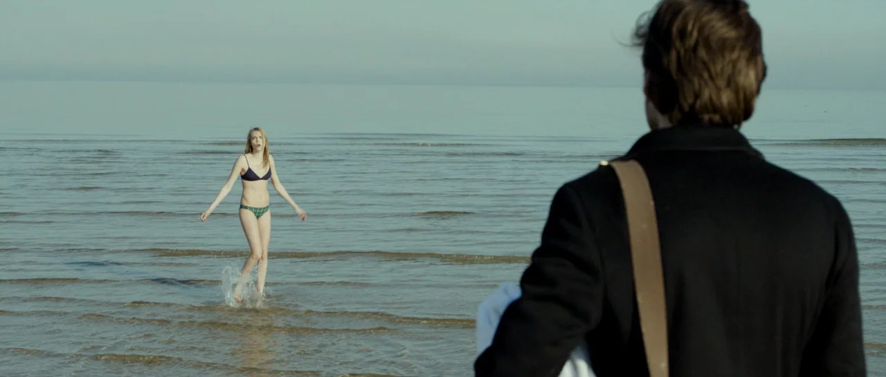 The Face of an Angel (2014) movie still: wide — A vast, calm shoreline meets a pale sky with gentle waves washing the sand. A woman wading through…