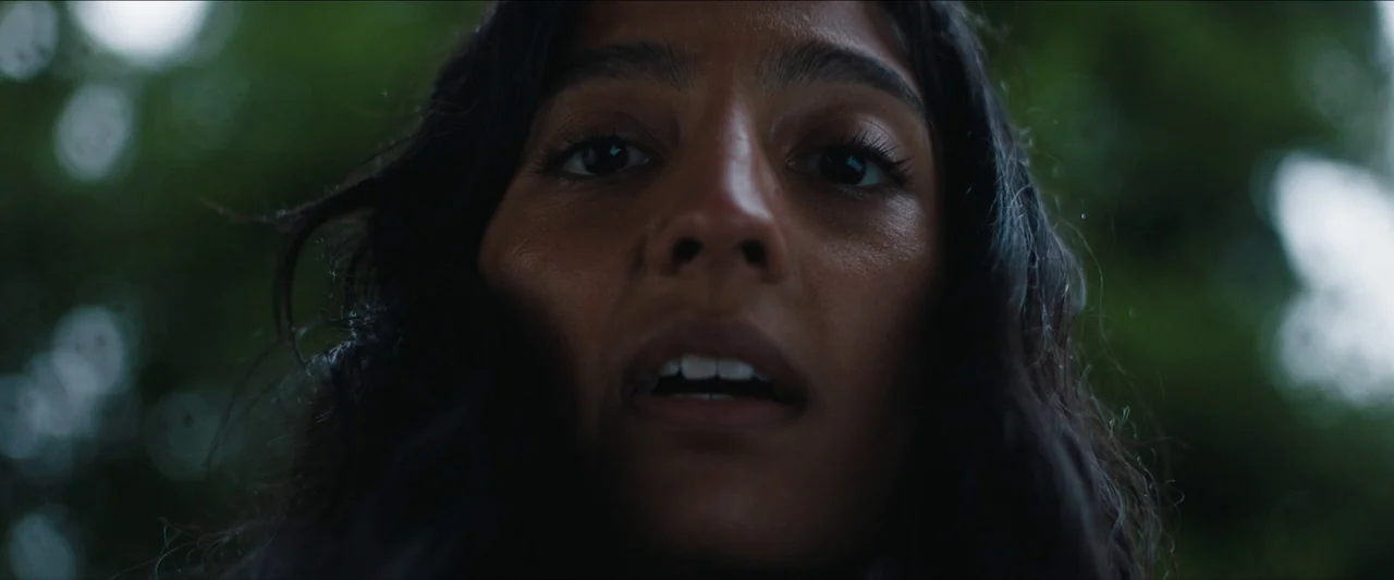 In The Earth (2021) movie still: closeup — The subject is framed from a high angle against a blurred green leafy background. She looks directly down…