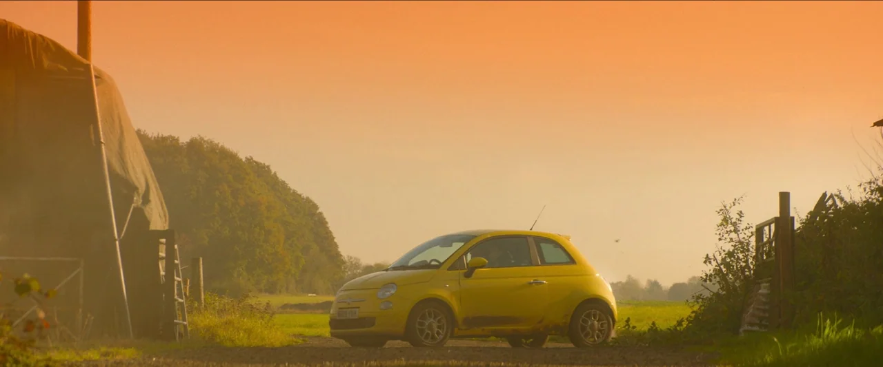 Unwelcome (2022) movie still: wide — A yellow compact car is parked on a dirt path next to a field. A large, dark-tarped structure stands on the…