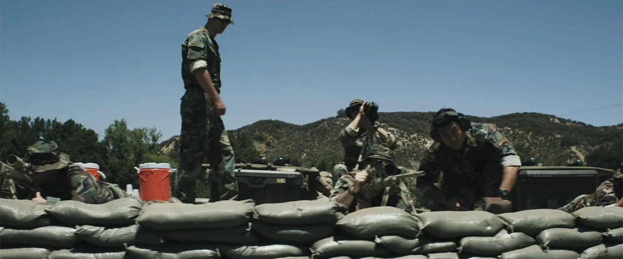 American Sniper (2014) movie still: wide — A group of soldiers stands behind a low sandbag wall on a dry hillside. The soldiers observe the terrain…