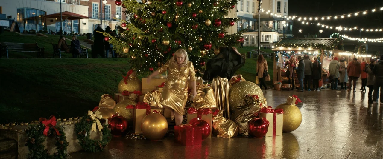 The Hustle (2019) movie still: medium — A public square is decorated with a large Christmas tree and numerous oversized golden ornaments. A woman in…