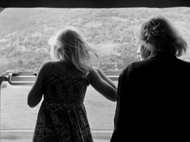 Alice in the Cities (1974) movie still: medium — Two individuals stand on the deck of a moving ferry looking toward the shore. A rolling hillside…