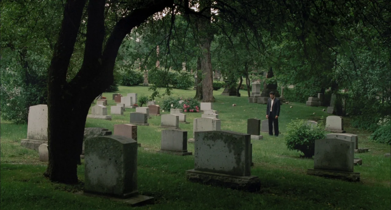 Incendies movie still: wide — A quiet cemetery is filled with numerous stone markers and green grass under trees. A lone man in a dark suit stands…