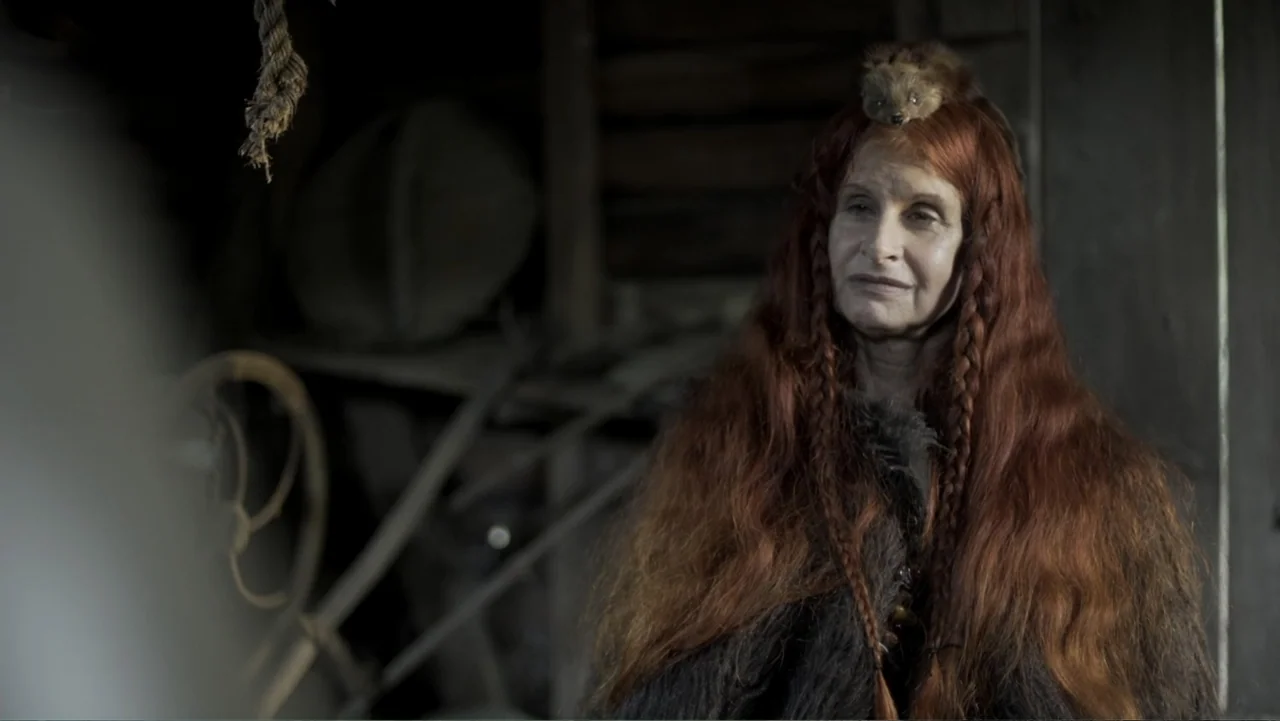 Kill Cavalry (2021) movie still: medium — A dark, wood-paneled interior space contains shadows and indistinct cluttered tools. An elderly woman with…