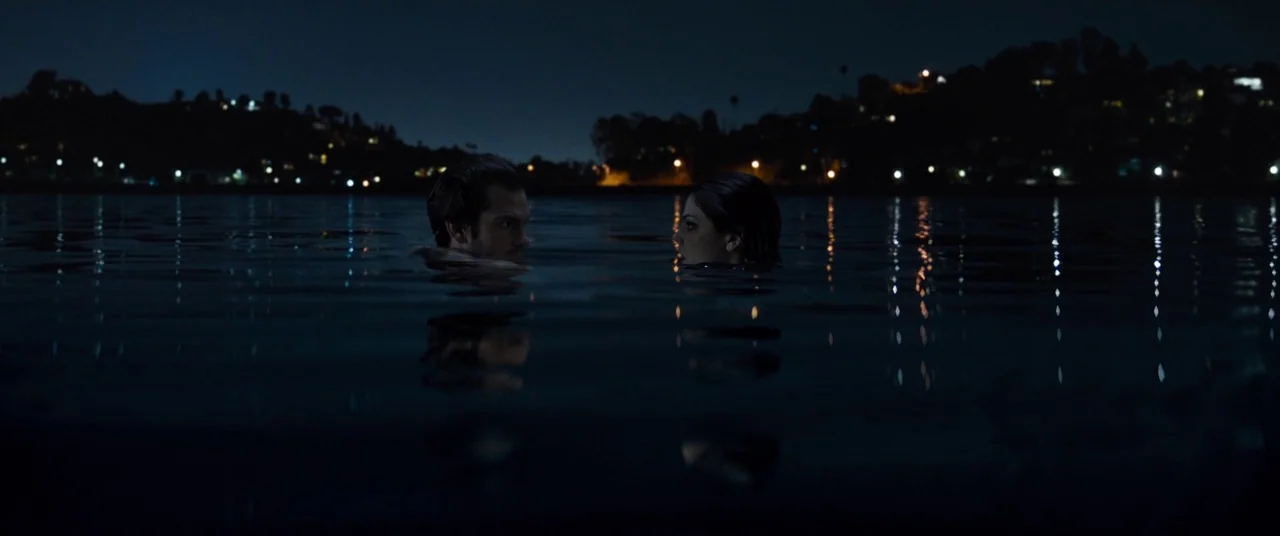 Under The Silver Lake (2018) movie still: wide — A wide shot shows a couple floating in a dark lake at night. Distant city lights illuminate the…