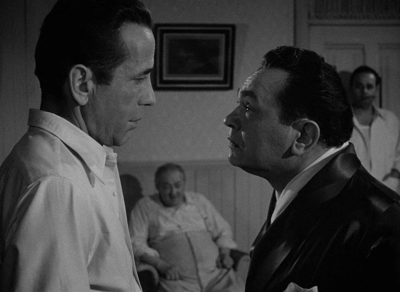 Key Largo (1948) movie still: medium — A dimly lit room with a framed picture on the wall above a couch. Two men face each other in the foreground…