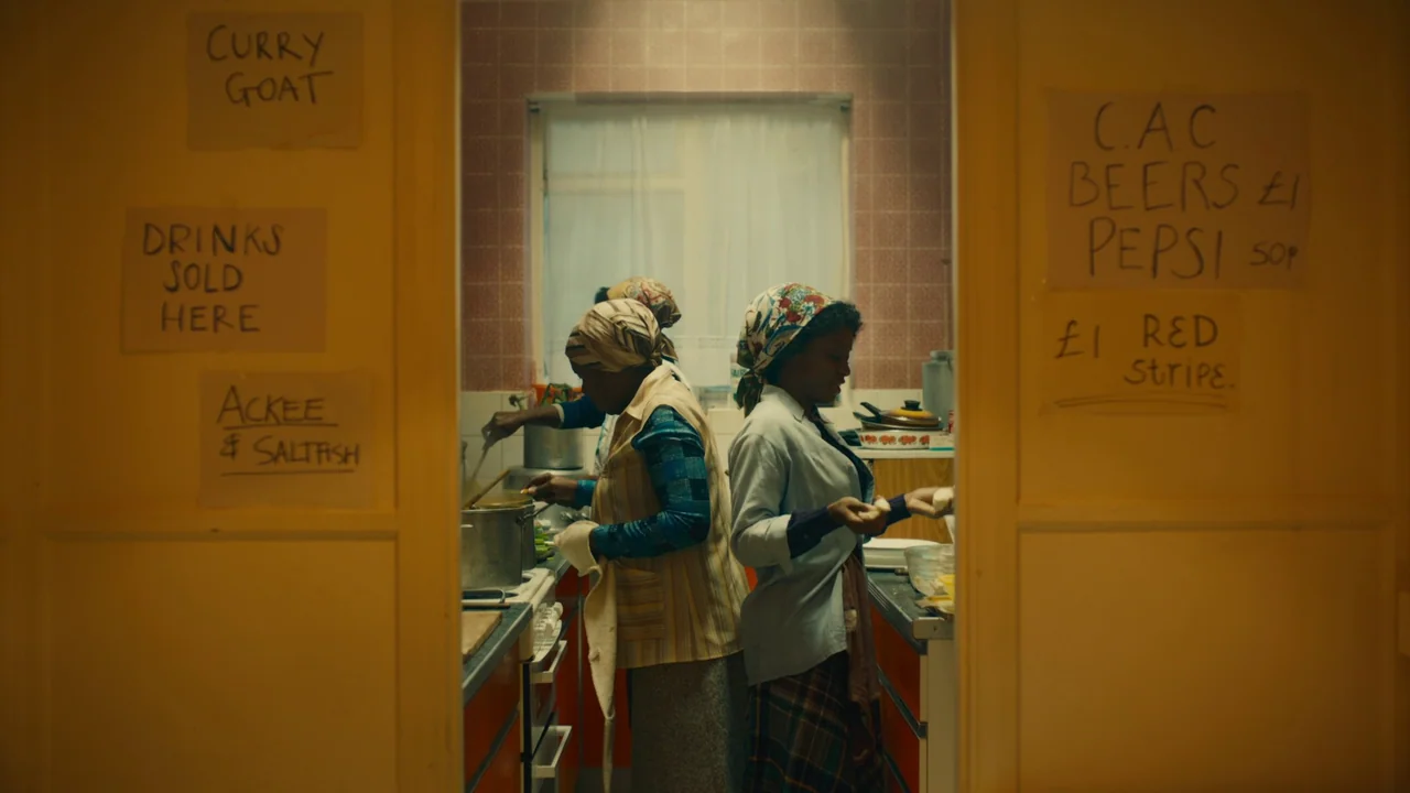 Lover’s Rock (2020) movie still: medium — A domestic kitchen with tiled walls and handwritten menu signs on the doorway frame. Two women work on…