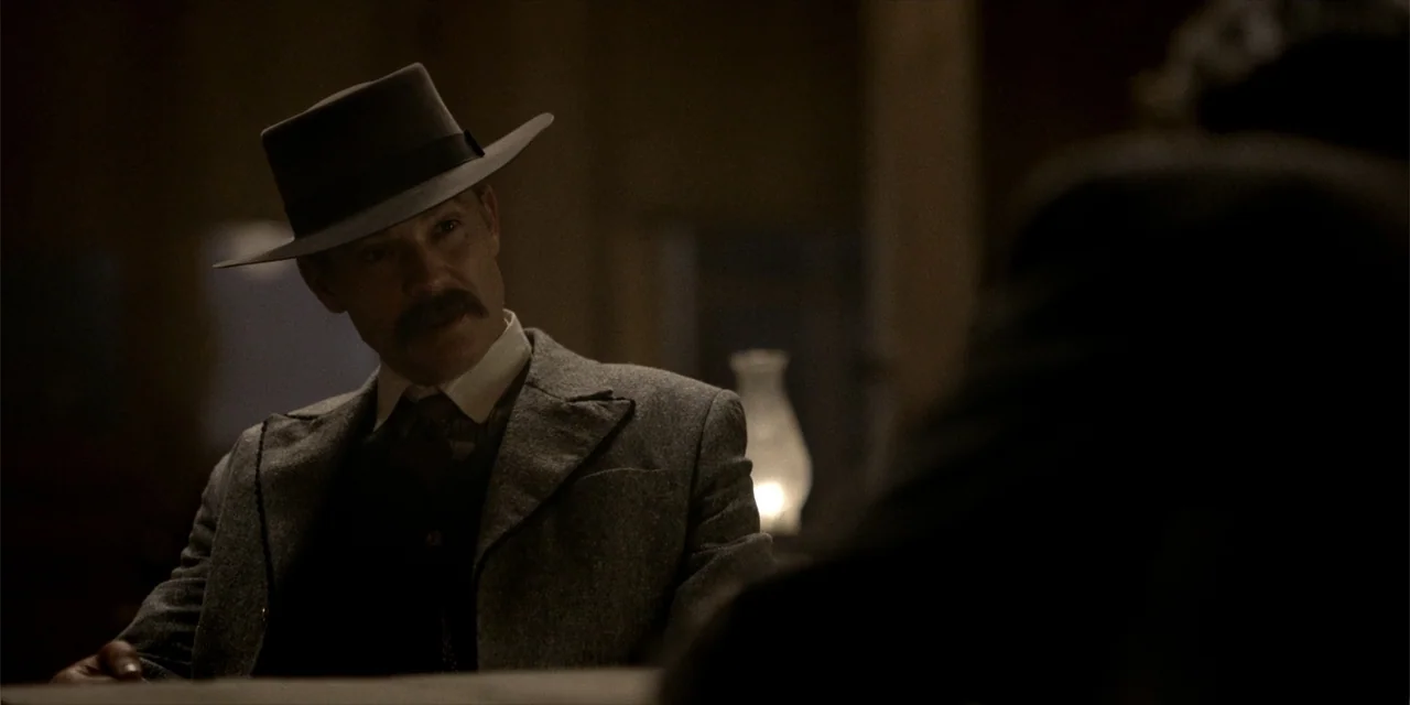 Deadwood: The Movie (2019) movie still: medium — A dimly lit room features a man sitting at a wooden table. The man wears a period-appropriate coat…