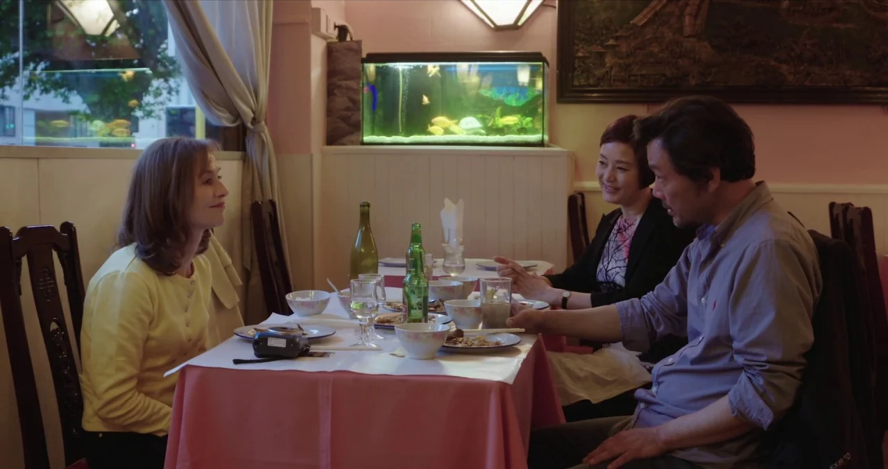 Claire’s Camera (2017) movie still: medium — A small dining area features pink tablecloths, dark wood chairs, and a lit fish tank. Three adults sit…